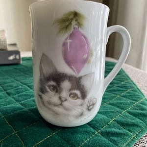 May include: White ceramic mug featuring a detailed illustration of a kitten with a Christmas ornament. The kitten has black and white fur and green eyes. A purple ornament with a green sprig is above the kitten. The mug has a curved handle.