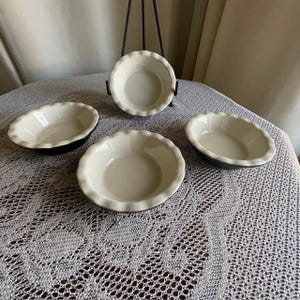 May include: Four small white ceramic pie dishes with fluted edges and black rims. The dishes are arranged on a white crocheted tablecloth.