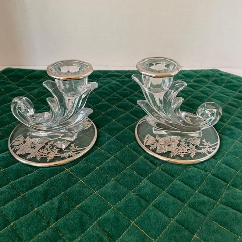 May include: Two clear glass candle holders with silver rims and floral designs. The candle holders have a unique, curved design with a silver base and a clear glass cup to hold the candle. The base has a silver floral design.