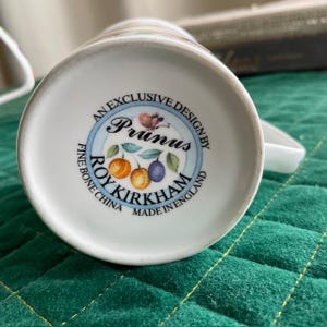 May include: White ceramic mug base with a circular design featuring the text "Prunus" and "Roy Kirkham Fine Bone China Made in England." The design includes illustrations of plums and oranges. The mug has a handle.