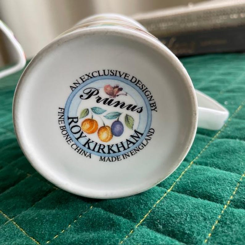 May include: White ceramic mug base with a circular design featuring the text "Prunus" and "Roy Kirkham Fine Bone China Made in England." The design includes illustrations of plums and oranges. The mug has a handle.