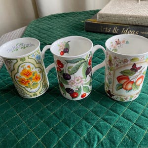May include: Three white ceramic mugs with floral and fruit designs. One mug features orange flowers and the text "The Herb Garden." Another has cherries and leaves, and the third has peaches and a butterfly. Each mug has a white handle.
