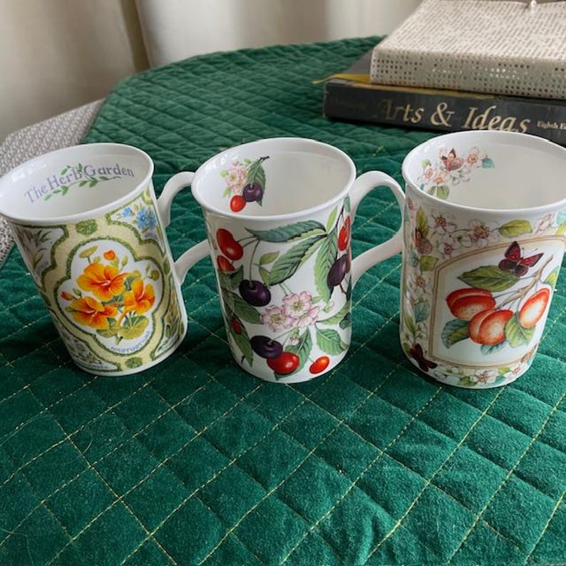 May include: Three white ceramic mugs with floral and fruit designs. One mug features orange flowers and the text "The Herb Garden." Another has cherries and leaves, and the third has peaches and a butterfly. Each mug has a white handle.