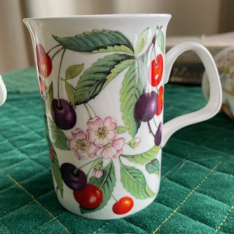 May include: White ceramic mug with a curved handle, adorned with a vibrant cherry design. The illustration features red and purple cherries, green leaves, and delicate pink blossoms. The mug is ideal for enjoying hot beverages.