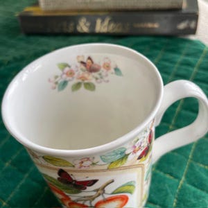May include: White ceramic mug with a curved rim and a white handle. The mug is decorated with a floral and butterfly design, featuring red apples and butterflies. The mug has a capacity of approximately 12 ounces.