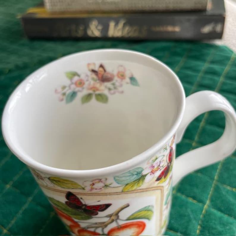 May include: White ceramic mug with a curved rim and a white handle. The mug is decorated with a floral and butterfly design, featuring red apples and butterflies. The mug has a capacity of approximately 12 ounces.