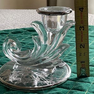 May include: A clear glass candle holder with a floral design on the base and a decorative, curved shape. The candle holder is approximately 4 inches tall. The base has a silver rim and floral etching.