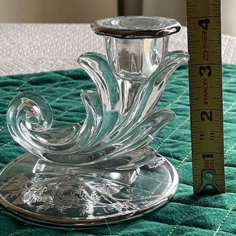 May include: A clear glass candle holder with a floral design on the base and a decorative, curved shape. The candle holder is approximately 4 inches tall. The base has a silver rim and floral etching.