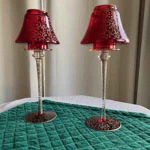 May include: Two red glass candle holders with gold leaf designs. The candle holders have a tall, slender stem and a wide, round base.