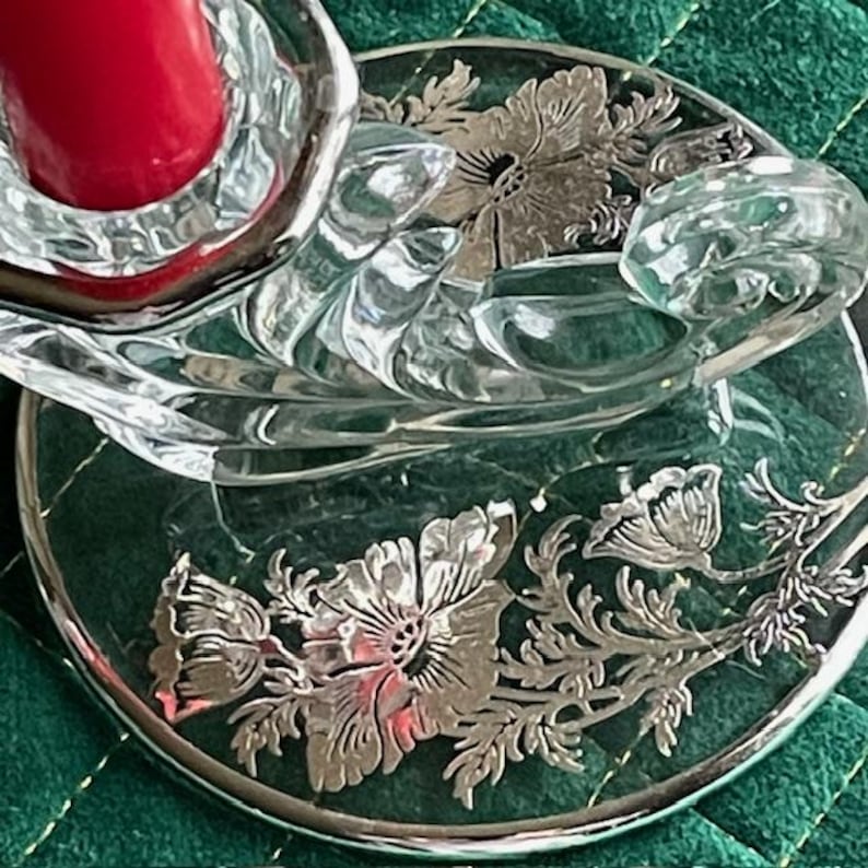 May include: A clear glass candle holder with a red candle. The holder has a floral design in silver. The candle holder sits on a green surface. The candle holder is a decorative item.