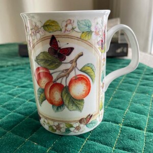 May include: White ceramic mug with a handle, featuring a detailed illustration of red cherries, a butterfly, and floral accents. The design is framed by a decorative border. The mug is suitable for hot beverages.