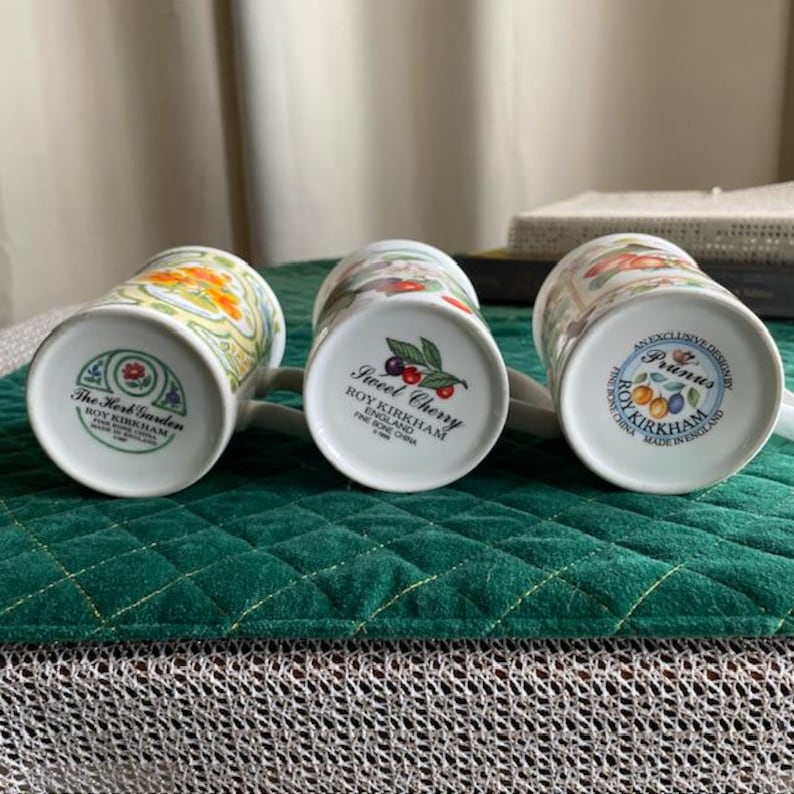 May include: Three white ceramic mugs with floral and fruit designs. The mugs are labeled "The Herb Garden", "Sweet Cherry", and "Prunus". Each mug has a white handle and is set on a green quilted surface.