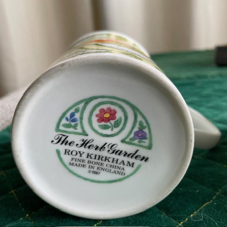 May include: White ceramic mug with a floral design and the text "The Herb Garden." The bottom of the mug features a decorative emblem with flowers and the text "ROY KIRKHAM FINE BONE CHINA MADE IN ENGLAND &copy;1997."