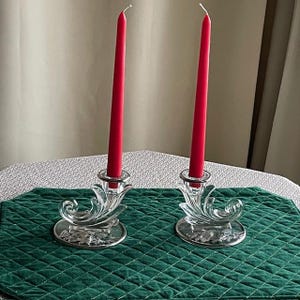 May include: Two clear glass candle holders with red taper candles. The candle holders have a decorative, swirling design and sit on a silver-colored base. The candles are tall and slender, adding a touch of elegance.