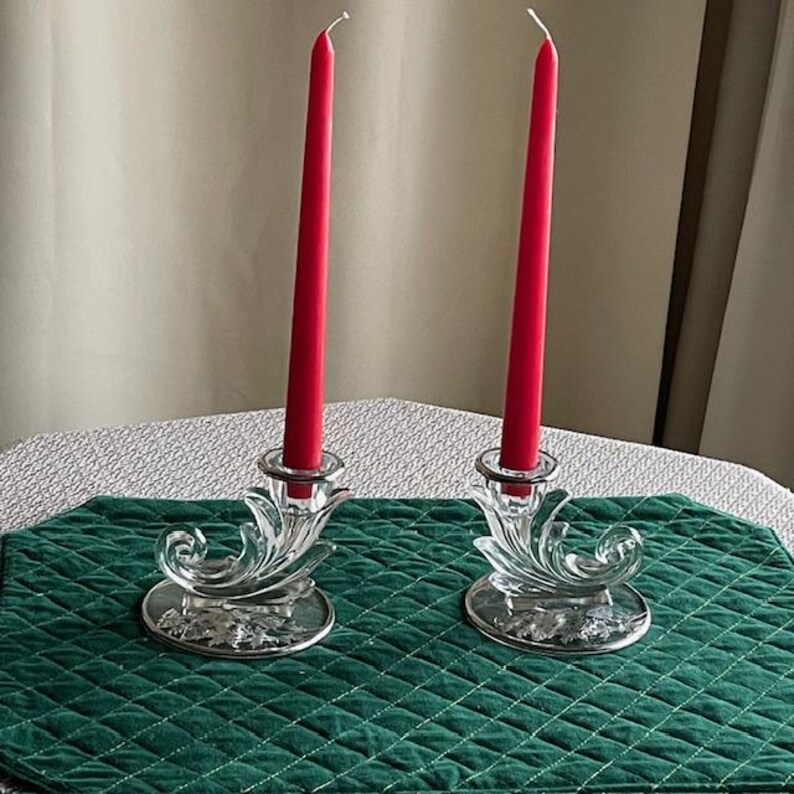May include: Two clear glass candle holders with red taper candles. The candle holders have a decorative, swirling design and sit on a silver-colored base. The candles are tall and slender, adding a touch of elegance.