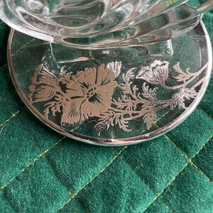 May include: A clear glass dish with a silver floral design on the base. The base is round with a silver rim and features a detailed flower and leaf pattern. The dish sits on a green quilted surface.