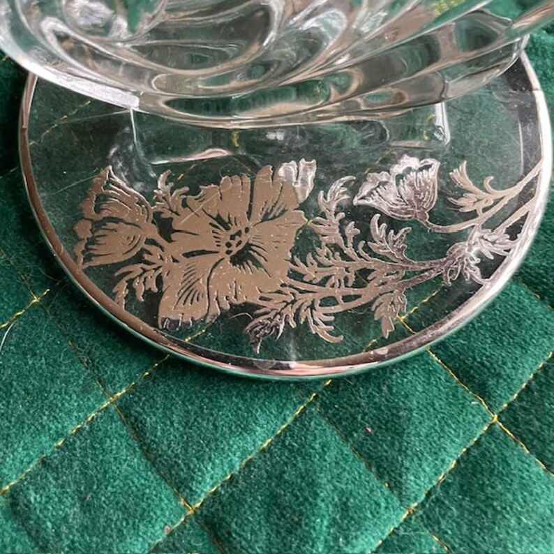 May include: A clear glass dish with a silver floral design on the base. The base is round with a silver rim and features a detailed flower and leaf pattern. The dish sits on a green quilted surface.