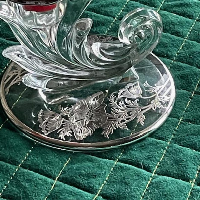 May include: A clear glass candle holder with a floral design. The base has a silver rim and features etched flowers and foliage. The candle holder has a curved, flowing design and sits on a green surface.