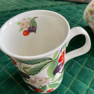 May include: White ceramic mug with a curved handle, decorated with a cherry blossom design. The design includes red and purple cherries, green leaves, and pink flowers. The mug has a wide opening and a smooth, glossy finish.