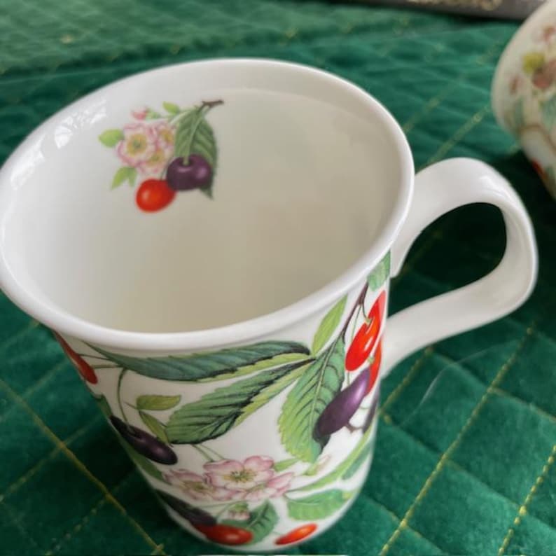 May include: White ceramic mug with a curved handle, decorated with a cherry blossom design. The design includes red and purple cherries, green leaves, and pink flowers. The mug has a wide opening and a smooth, glossy finish.