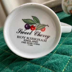 May include: White fine bone china teacup base with a "Sweet Cherry" design. The design includes a cluster of cherries and leaves. The text "ROY KIRKHAM ENGLAND FINE BONE CHINA &copy;1999" is printed below the design.