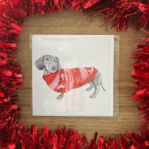 以下が含まれることがあります： 赤いクリスマスセーターを着たダックスフンドの鉛筆画が描かれたグリーティングカードの束。カードは木製の表面にあり、赤いガーランドで囲まれています。