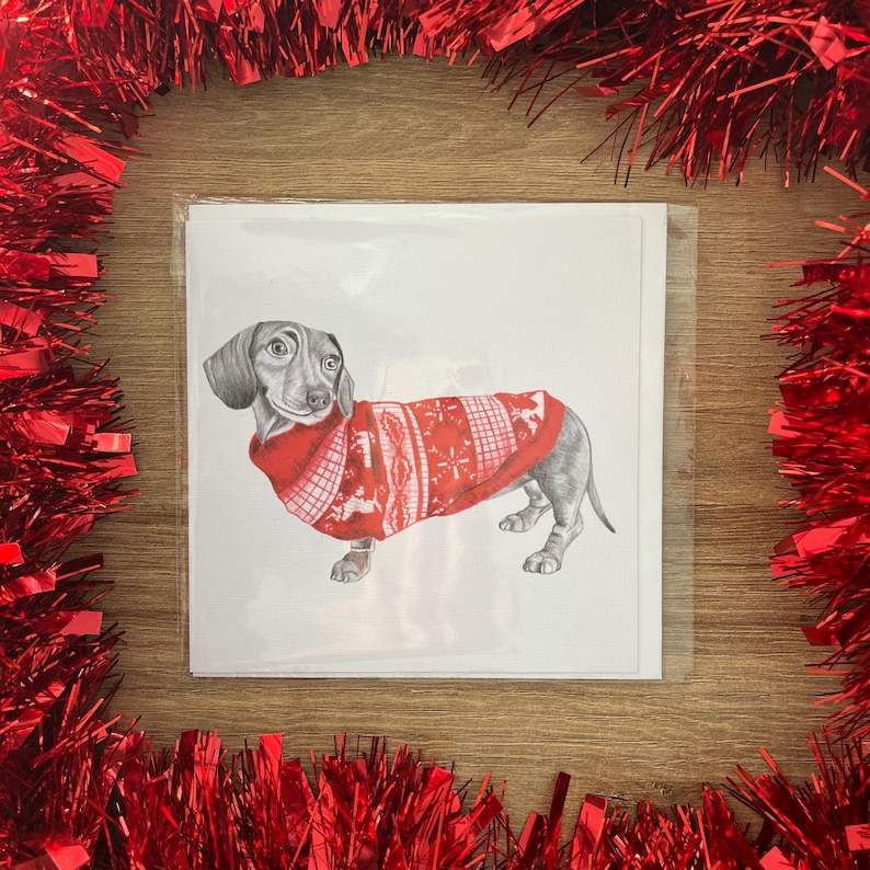 以下が含まれることがあります： 赤いクリスマスセーターを着たダックスフンドの鉛筆画が描かれたグリーティングカードの束。カードは木製の表面にあり、赤いガーランドで囲まれています。