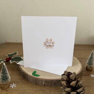 以下が含まれることがあります： 「Aimee's Pawtraits」のテキストと足跡のロゴが入った白いグリーティングカード。クリスマスデコレーションが施された木の輪切りに飾られています。松ぼっくり、小さなクリスマスツリー、ヒイラギも写っています。