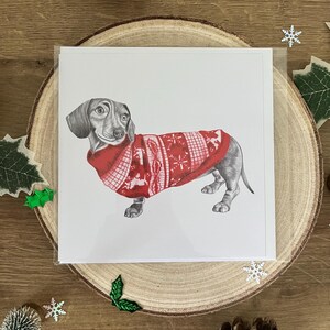 以下が含まれることがあります： 赤いクリスマスセーターを着たダックスフンドの詳細なイラストが描かれたグリーティングカード。カードは木の輪切りに置かれ、松ぼっくりやヒイラギなどの装飾要素に囲まれています。