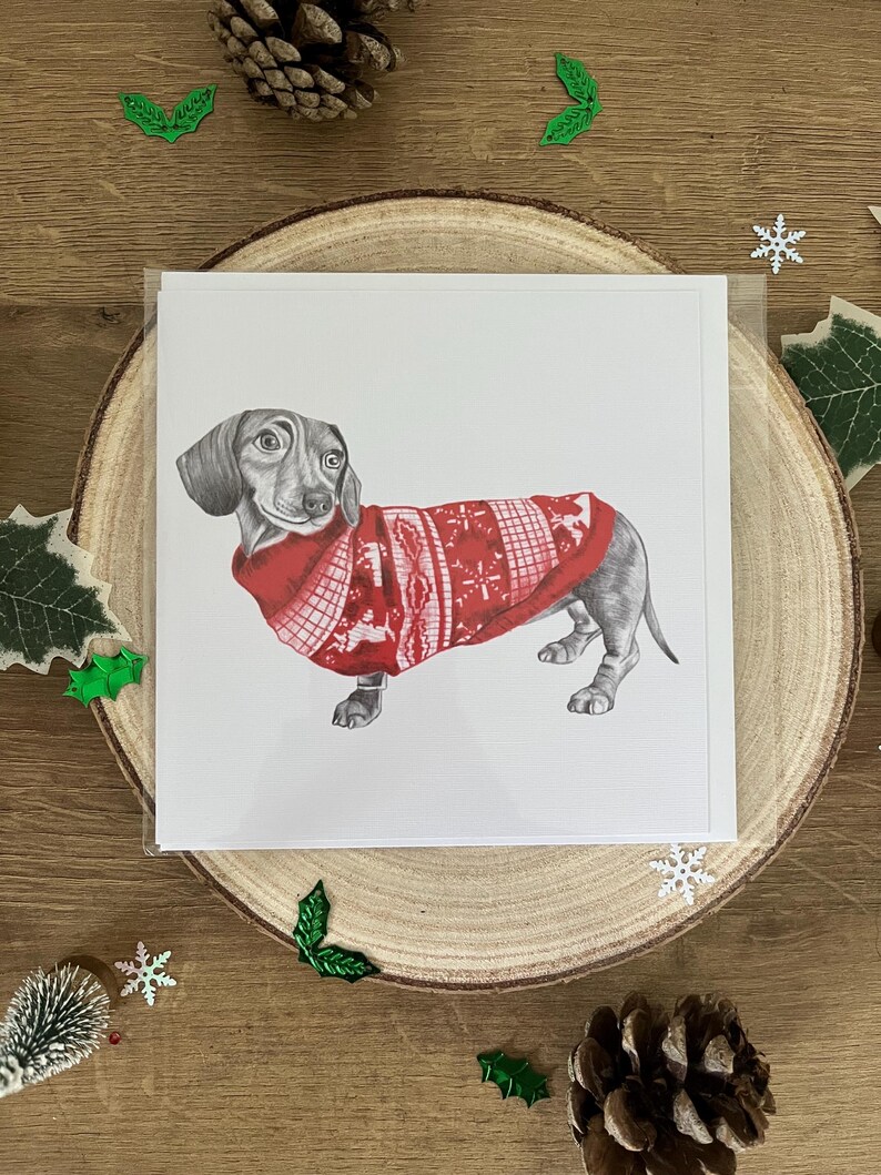 以下が含まれることがあります： 赤いクリスマスセーターを着たダックスフンドの詳細なイラストが描かれたグリーティングカード。カードは木の輪切りに置かれ、松ぼっくりやヒイラギなどの装飾要素に囲まれています。