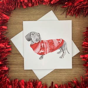 以下が含まれることがあります： 白い模様の赤いクリスマスセーターを着たダックスフンドの鉛筆画。カードは白い封筒の上にあり、木製の表面に赤いガーランドで囲まれています。