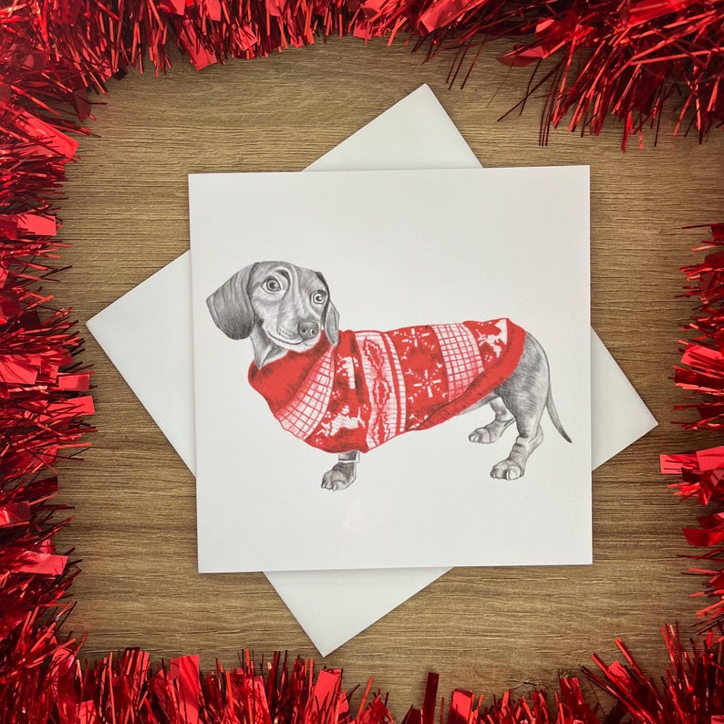 以下が含まれることがあります： 白い模様の赤いクリスマスセーターを着たダックスフンドの鉛筆画。カードは白い封筒の上にあり、木製の表面に赤いガーランドで囲まれています。