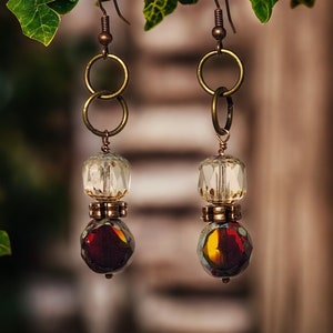 Puede incluir: Un par de pendientes de tono dorado con cuentas de vidrio transparentes y rojas. Los pendientes tienen un diseño de doble anillo y cuelgan de un gancho.