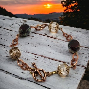 May include: A copper chain bracelet with a toggle clasp. The bracelet features four glass beads, two with a clear, speckled design and two with a dark brown, speckled design.