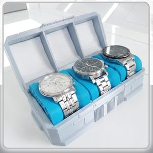 May include: A light gray, rectangular watch organizer box with three compartments, each containing a bright blue watch pillow. Three silver-toned metal wristwatches with round faces and link bracelets are displayed on the pillows. Text reads WATCH ORGANIZER BOX.