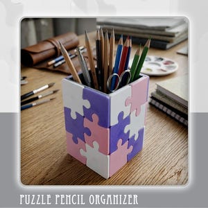 Puede incluir: Un organizador de lápices en forma de rompecabezas en blanco, rosa y morado, que contiene lápices y unas tijeras. El texto "PUZZLE PENCIL ORGANIZER" y "Digital Files Only" se muestran debajo. Otros materiales de arte son visibles en el fondo.