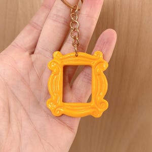 Könnte beinhalten: Ein leuchtend gelber Schlüsselanhänger in Form eines Bilderrahmens mit einer goldfarbenen Kette und einem Schlüsselring. Der Rahmen hat ein dekoratives, verziertes Design. Der Schlüsselanhänger wird von einer Hand gehalten.