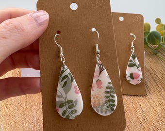 May include: Teardrop-shaped earrings with a floral pattern of pink, green, and beige on a white background. The earrings hang from silver-colored hooks and are displayed on brown cardstock.