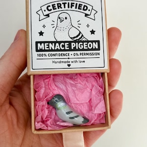 May include: A small grey and black pigeon figurine nestled inside a brown cardboard matchbox lined with pink tissue paper. The box lid features a black and white illustration of a pigeon with the text CERTIFIED MENACE PIGEON 100% CONFIDENCE 0% PERMISSION.