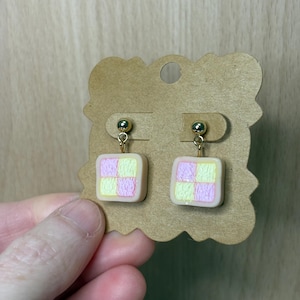 Op de afbeelding: Een paar handgemaakte oorbellen met een vierkant ontwerp en een roze en geel geblokt patroon. De oorbellen zijn bevestigd aan goudkleurige pinnen en hangen aan een kleine goudkleurige bal. De oorbellen worden gepresenteerd op een bruine kaart.