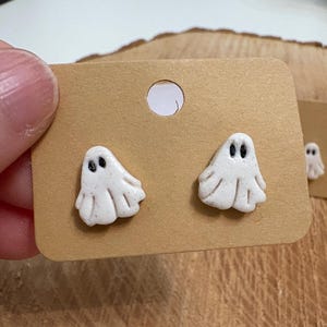 Può includere: Un paio di orecchini bianchi a forma di fantasma con occhi neri, montati su un cartoncino marrone per orecchini. Gli orecchini hanno un aspetto strutturato e sono progettati per assomigliare a fantasmi amichevoli. Il cartoncino ha un foro circolare vicino alla parte superiore.