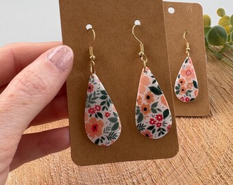 May include: Floral teardrop earrings with gold-colored hooks. The earrings feature a white background with a pattern of orange, red, and green flowers and leaves. The earrings are displayed on a brown card.