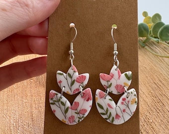 May include: Floral-patterned earrings with a white base and pink and yellow flower accents. The earrings are made of a lightweight material and feature silver-colored hooks. The design includes two overlapping petal shapes.