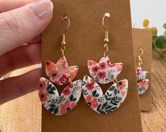 May include: Floral dangle earrings with gold-colored hooks. The earrings feature a white base with a pattern of red and peach flowers and green leaves. The earrings are displayed on a brown card.