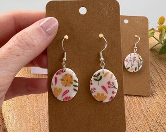 May include: Floral dangle earrings with a white background and colorful flowers in yellow, pink, and green. The earrings are displayed on a brown earring card. The earrings have silver hooks.