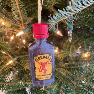May include: A miniature bottle ornament shaped like a Fireball Cinnamon Whisky bottle. The bottle is red, black, and gold with a red cap and a label that reads "Fireball Cinnamon Whisky, Red Hot, ALC 33% by Vol. 166 Proof, Flavored with Natural Cinnamon Flavor."