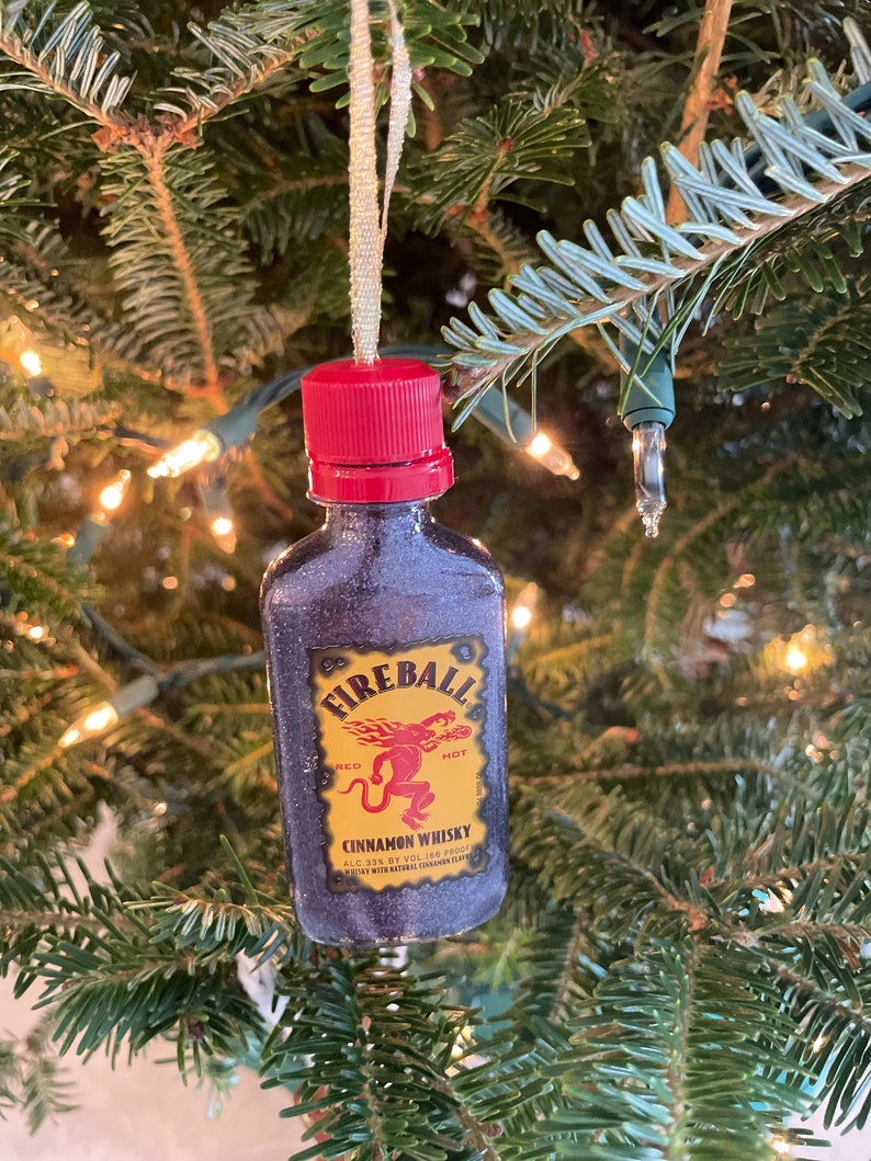 May include: A miniature bottle ornament shaped like a Fireball Cinnamon Whisky bottle. The bottle is red, black, and gold with a red cap and a label that reads "Fireball Cinnamon Whisky, Red Hot, ALC 33% by Vol. 166 Proof, Flavored with Natural Cinnamon Flavor."