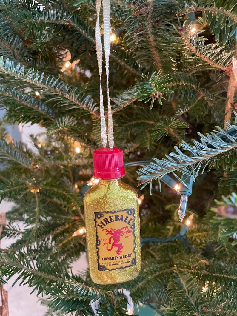 May include: Gold glitter Fireball Cinnamon Whisky bottle ornament with a red cap and a red dragon logo. The bottle is hanging on a Christmas tree.