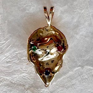 May include: A gold pendant with an irregular, textured surface. It has embedded gemstones in green, red, and blue. The pendant's bail is a V-shaped design.