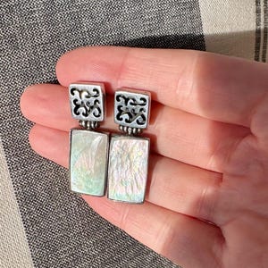 May include: A pair of silver-toned earrings. Each earring features a square top with an openwork design above a rectangular, iridescent, mother-of-pearl-like stone. The earrings are displayed against a striped background.